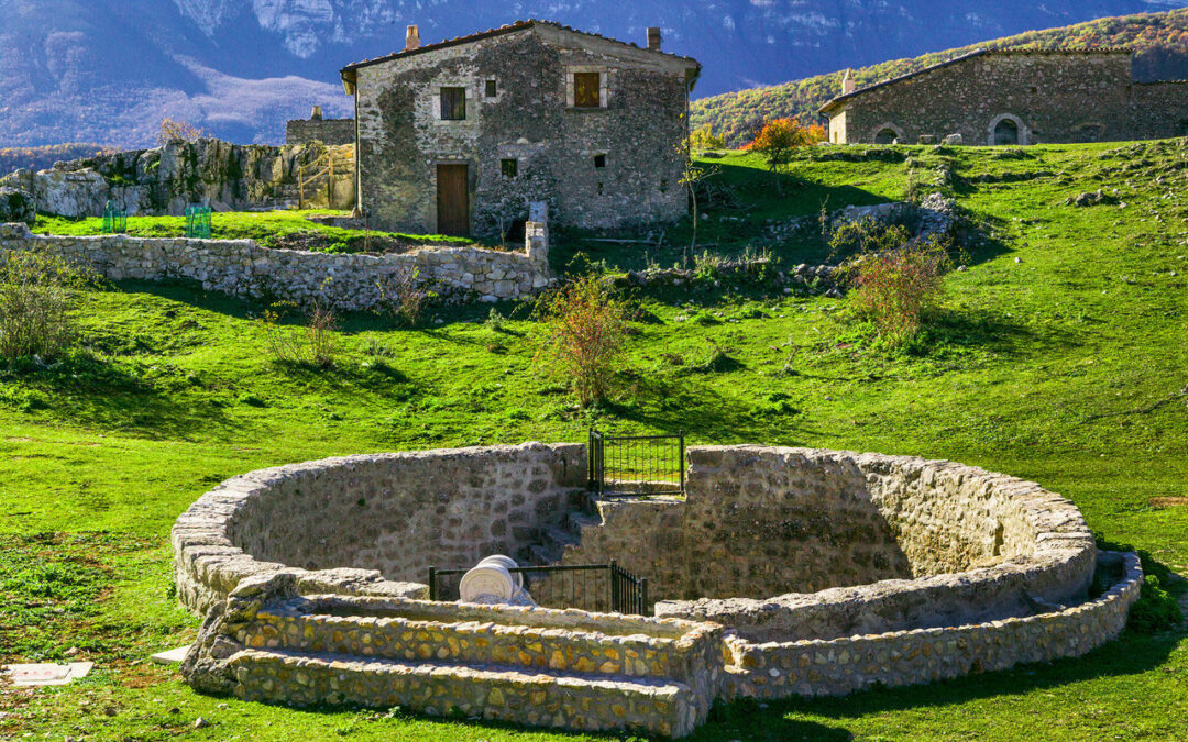 Vista delle Pagliare di Tione, antiche costruzioni in pietra immerse nel paesaggio montano abruzzese, con uno storico edificio e un antico recinto circolare in primo piano, circondati dal verde delle colline e dalle montagne sullo sfondo.