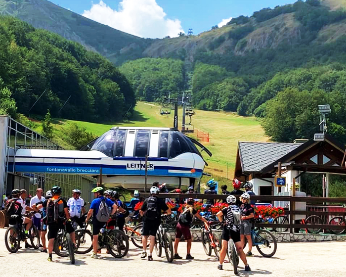 Gruppo di ciclisti pronti per un'escursione estiva a Campo Felice, vicino alla stazione della cabinovia Fontanavalle Brecciara, circondati da paesaggi verdi e montani.